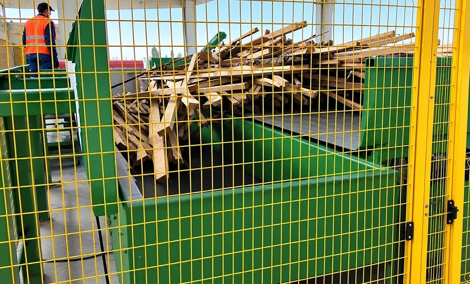Industrial wood waste shredder with green feed hopper, timber load, operator platform, and yellow safety guarding