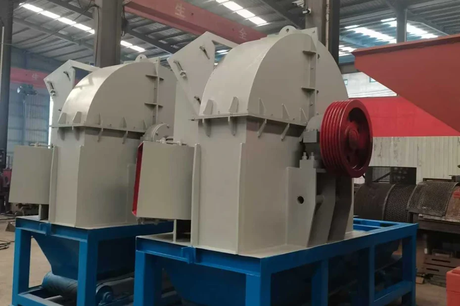 Enclosed drum wood chipper machine housing with belt drive guard and integrated discharge conveyor frame on a plant floor