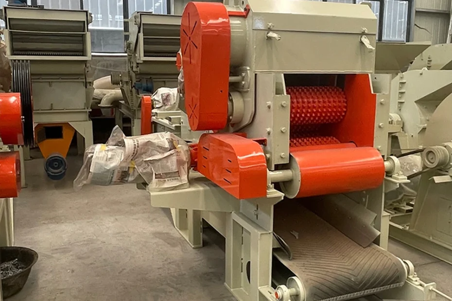 Industrial shredder with toothed crushing roller, orange safety guards, and integrated discharge conveyor in factory