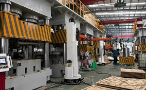 Heavy-duty four-column hydraulic press in factory assembly hall with striped safety guards and overhead crane